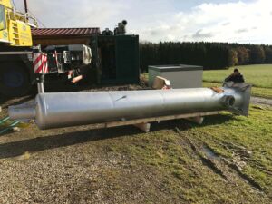Großes silbernes Abgasrohr auf Holzbalken, dahinter Kran und grüner Container