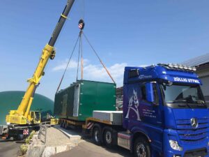 Kran hebt einen grünen BHKW-Container von einem blauen Lastwagen