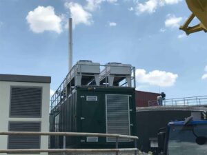 Grüner BHKW-Container mit Kühlern auf dem Dach neben einem Gebäude