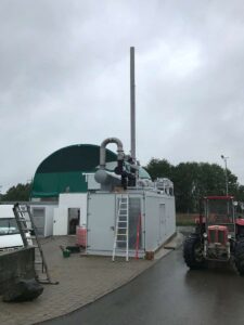 Grauer BHKW-Container mit Schornstein vor einer grünen Kuppel, daneben ein Traktor