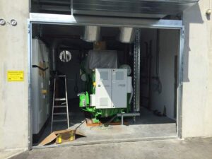 Blick durch ein offenes Tor auf ein grünes BHKW-Modul mit Schaltschrank