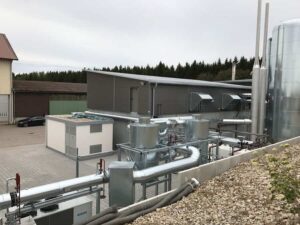 Isolierte Rohrleitungen zwischen weißen Technikcontainern vor einer Halle