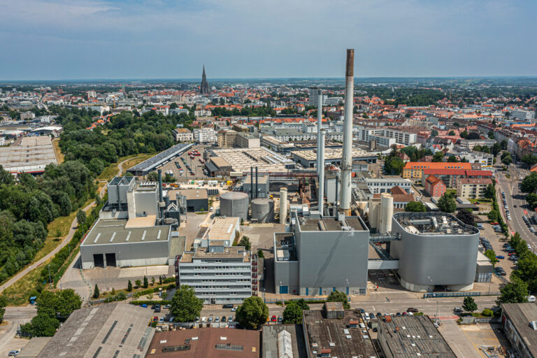 Luftaufnahme eines Heizkraftwerks mit hohem Schornstein am Rand einer Stadt