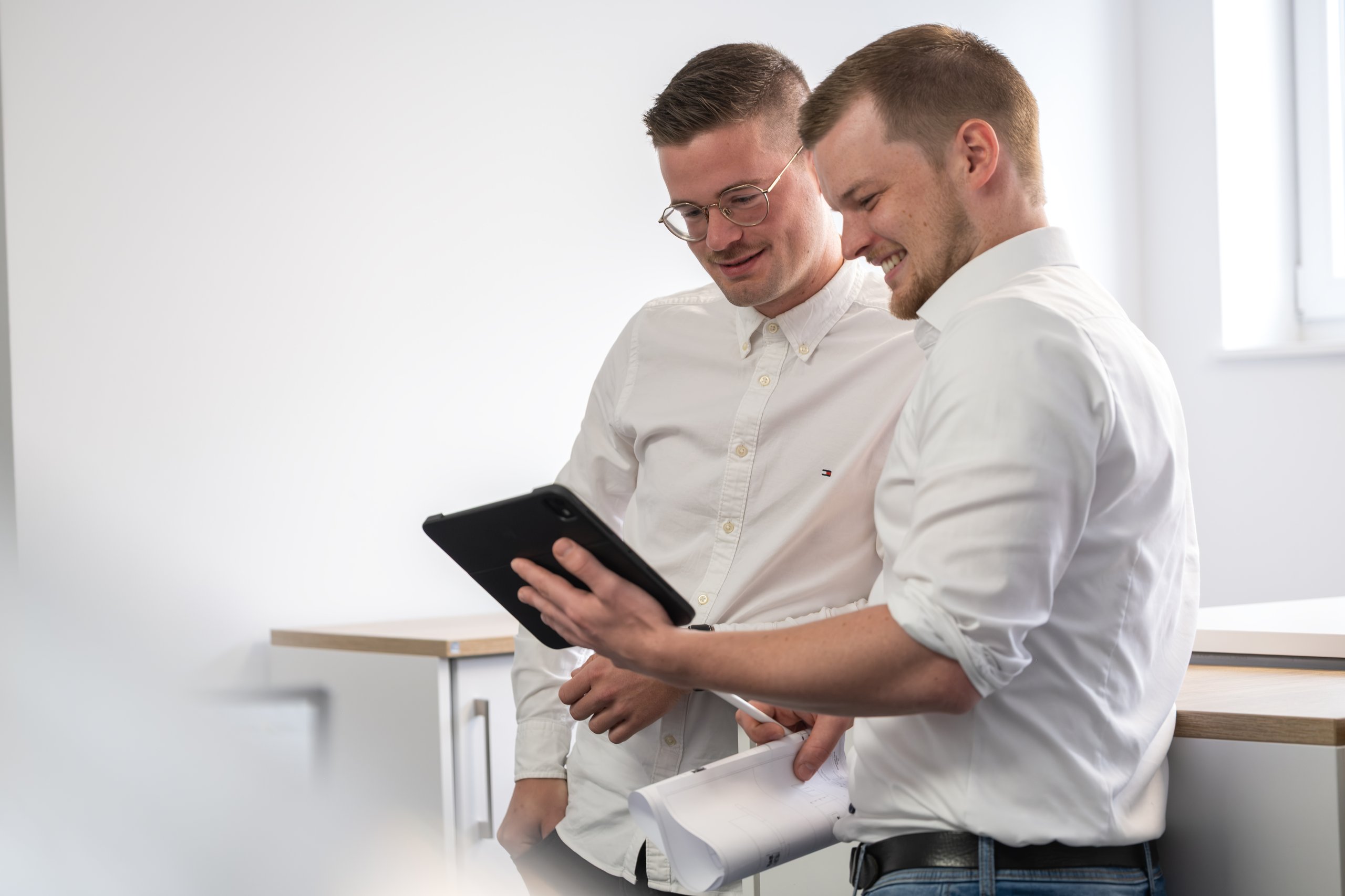 Zwei lächelnde Mitarbeiter in weißen Hemden schauen in einem hellen Raum gemeinsam auf ein Tablet
