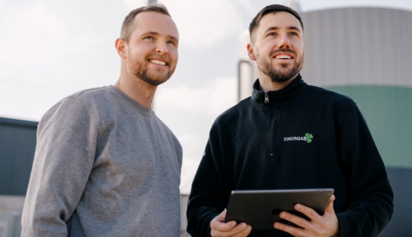 Zwei Mitarbeiter vor einer Biogasanlage, einer hält ein Tablet, im Hintergrund sind große Fermenter und Industrieanlagen für erneuerbare Energie zu sehen.