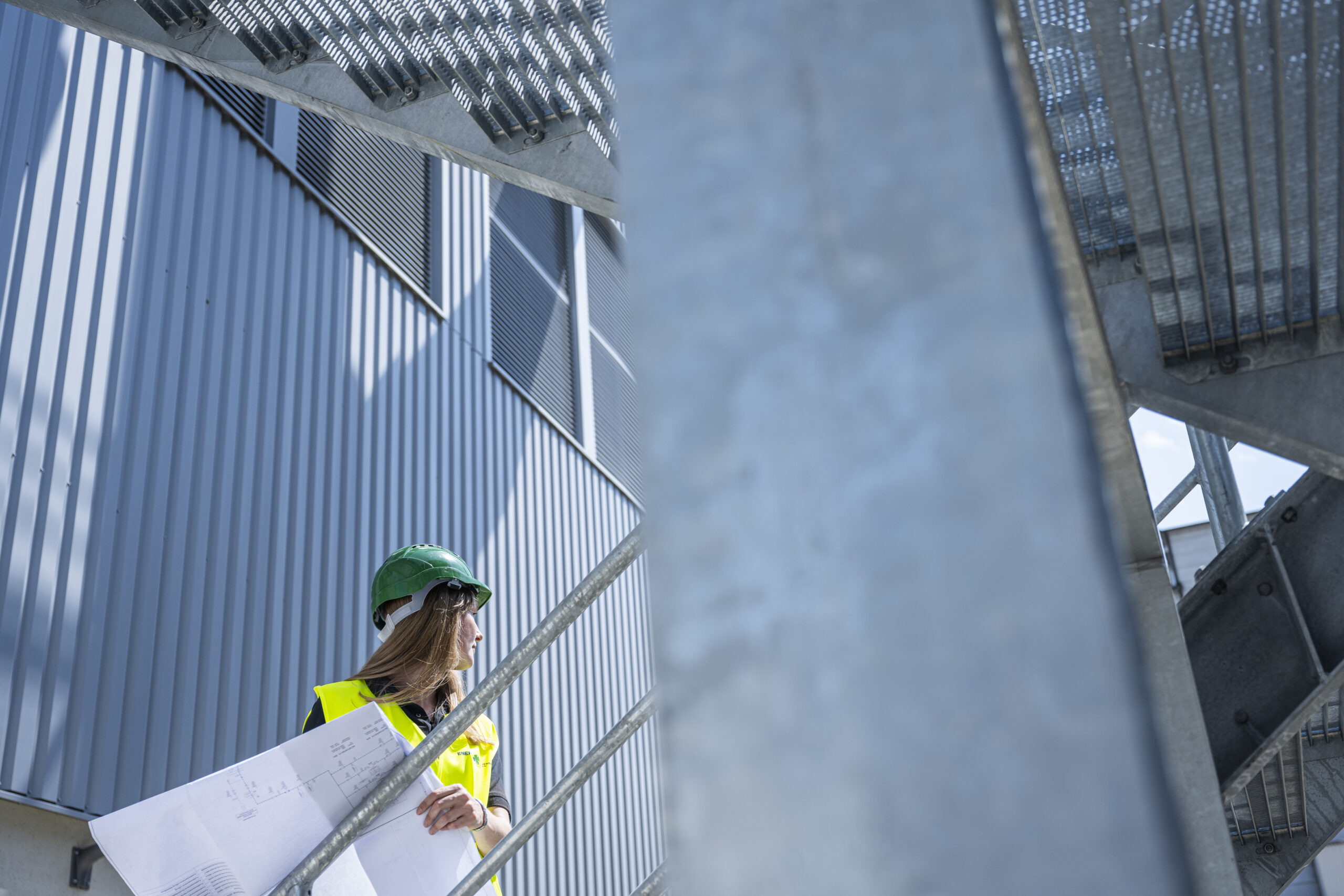 Frau mit Schutzhelm und Warnweste hält einen Plan auf einer Stahltreppe
