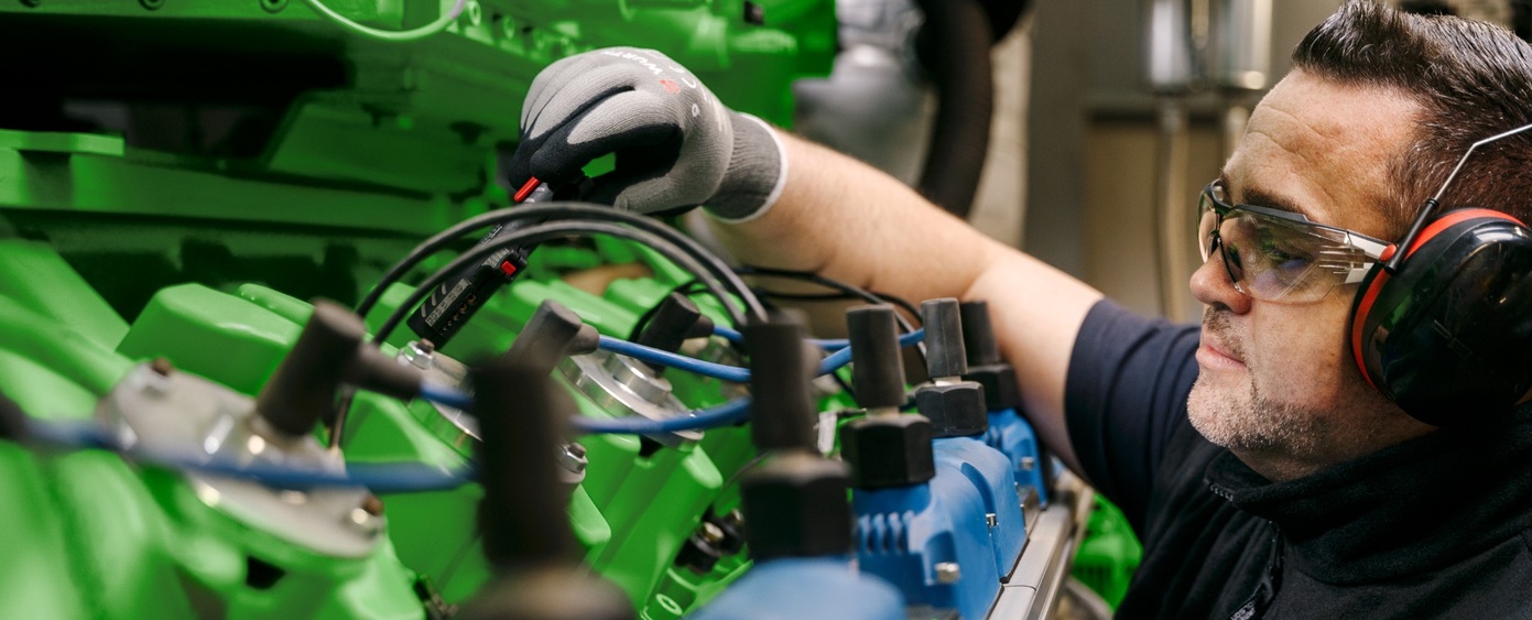 Mann mit Schutzbrille und Gehörschutz arbeitet an Kabeln eines grünen Motors