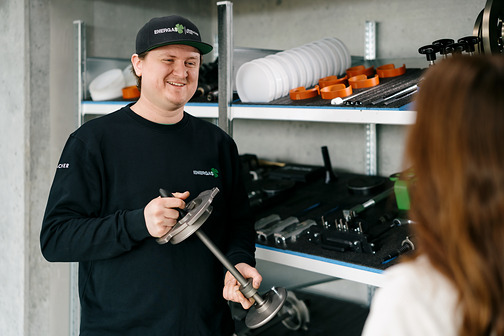 Lächelnder Mann mit Kappe hält ein Metallteil in einem Lagerraum