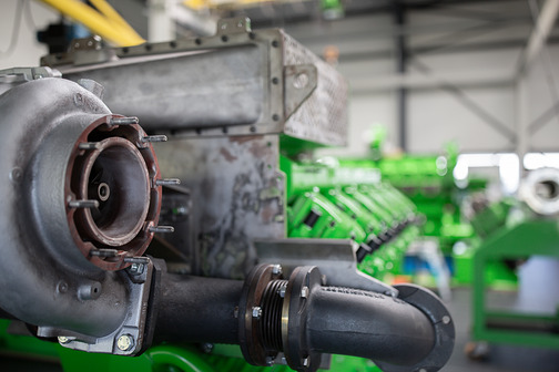 Nahaufnahme eines Turboladers mit Rohrleitung, dahinter unscharf grüne Motoren in der Schulungshalle