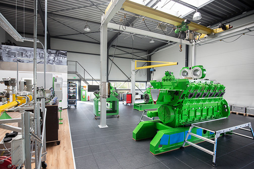 Blick in die Schulungshalle: großer grüner Motor mit Arbeitspodest, gelber Laufkran und links Übungsstände mit Rohrleitungen